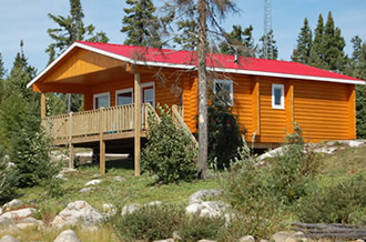 A small orange cabin with a red roof, surrounded by trees and rocks, under a clear blue sky.