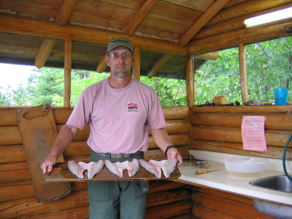 Man holding a tray with four fish fillets in a wooden kitchen with a sink, cutting board, and leafy background.