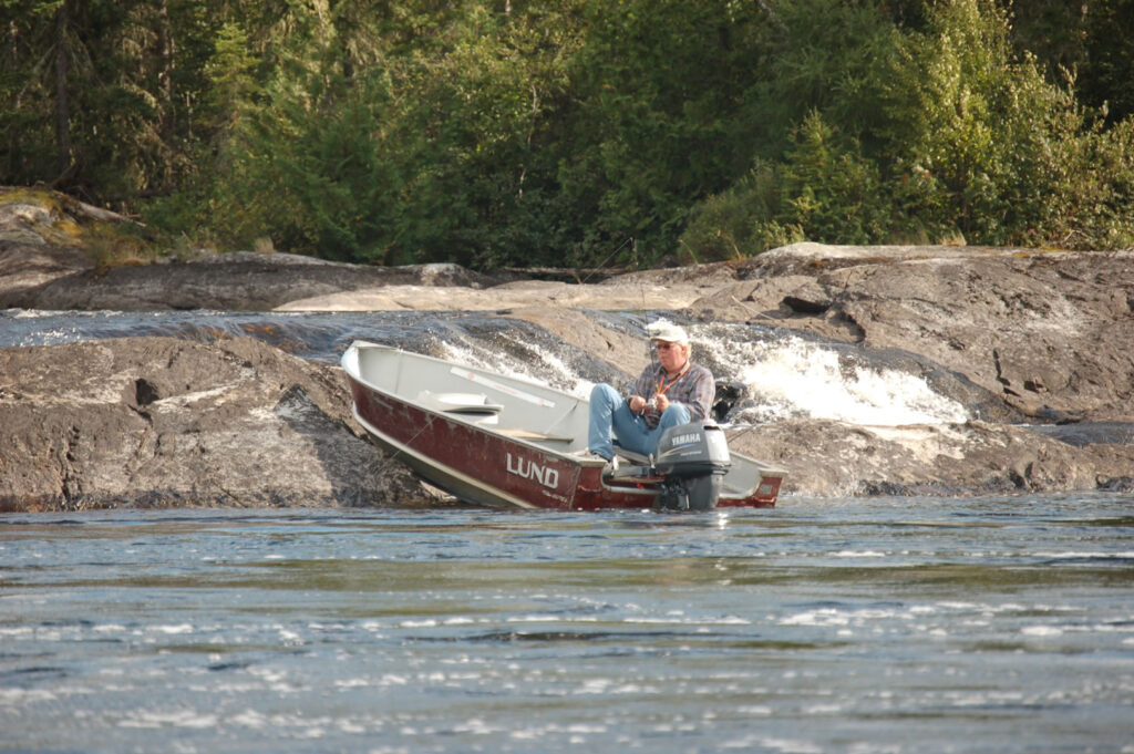 Man sitting in a small motorboat on rocky shore by a forested area, under clear skies.