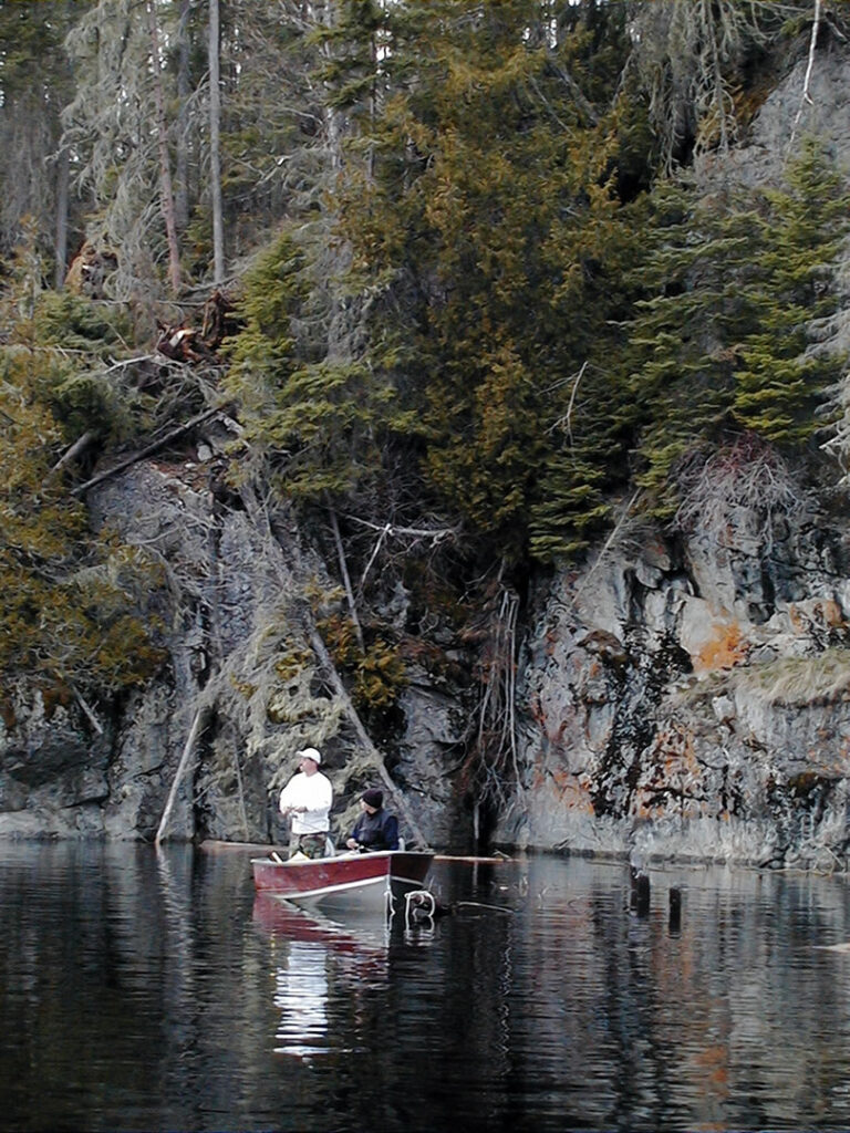 Two people in a small boat fishing on a calm lake, surrounded by tall trees and rocky cliffs.