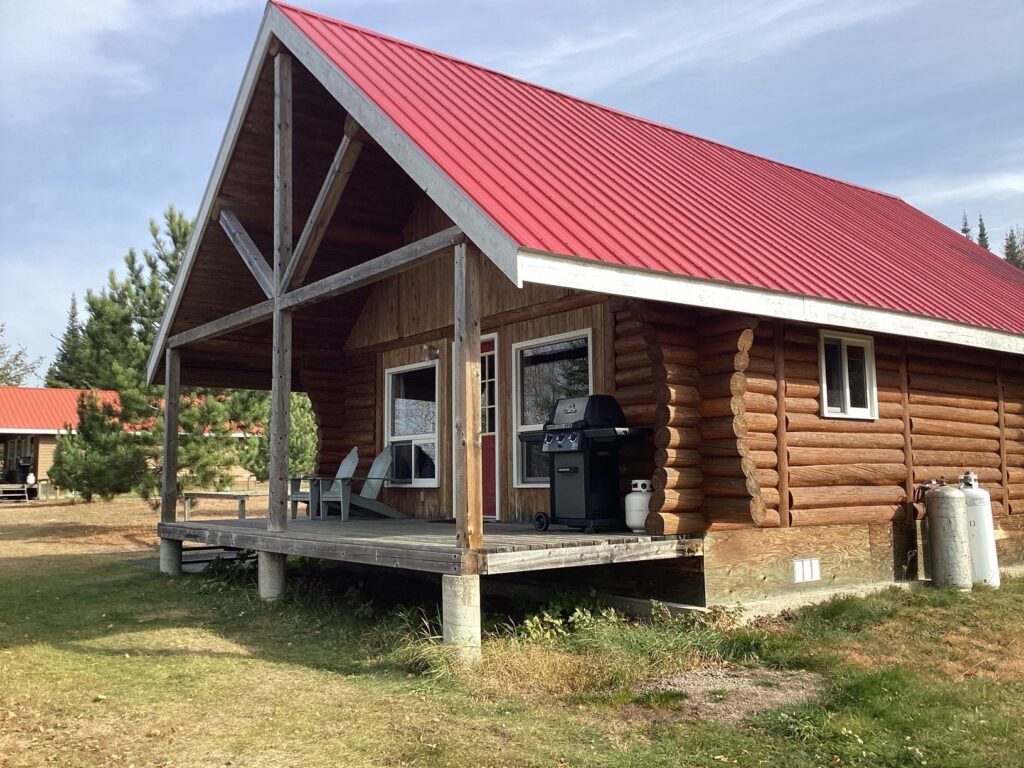 Log cabin with a red metal roof, front porch, two chairs, and a grill, set in a grassy area with trees in the background.