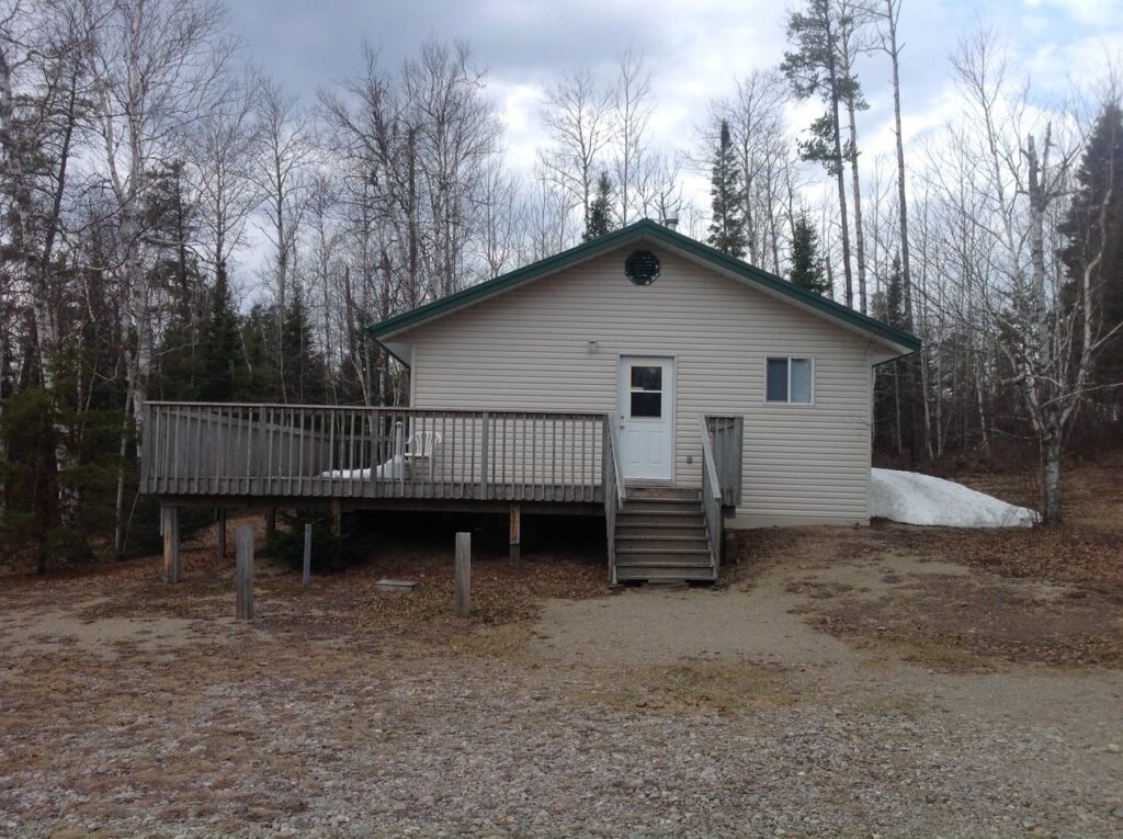 Small beige cabin with a green roof and wooden deck, surrounded by bare trees. Snow patches on the ground, overcast sky.