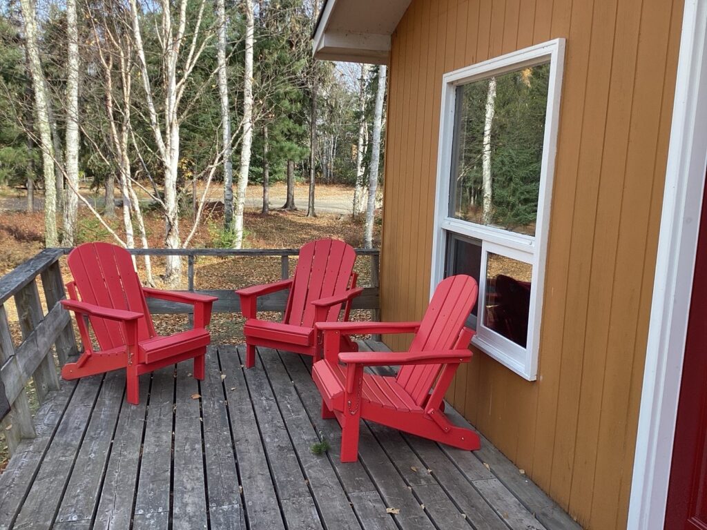 Three red Adirondack chairs on a wooden deck beside a brown cabin, with trees in the background.