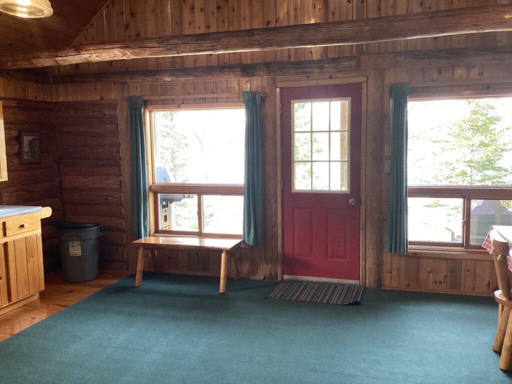 Interior of a wooden cabin with a red door, two windows with teal curtains, a green carpet, and a wooden bench.