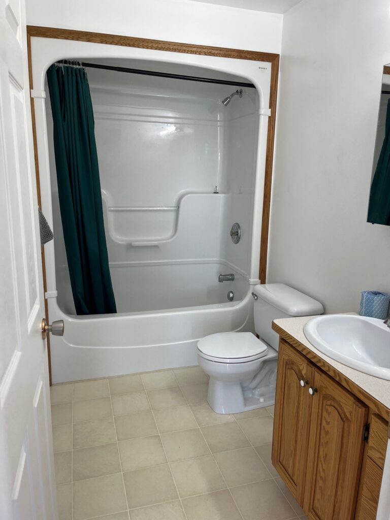 Bathroom with a white bathtub and shower, green curtain, toilet, and wooden vanity with a sink. White walls and tiled floor.