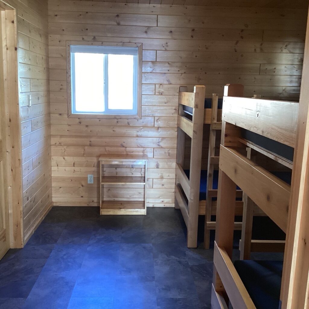 Wooden room with bunk beds, a small shelf, and a window. Walls and beds are made of light wood. Floor is dark tile.