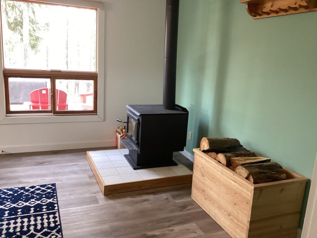 Wood stove on a tiled base with logs stored in a wooden box. Large window shows patio chairs outside. Room features green wall.