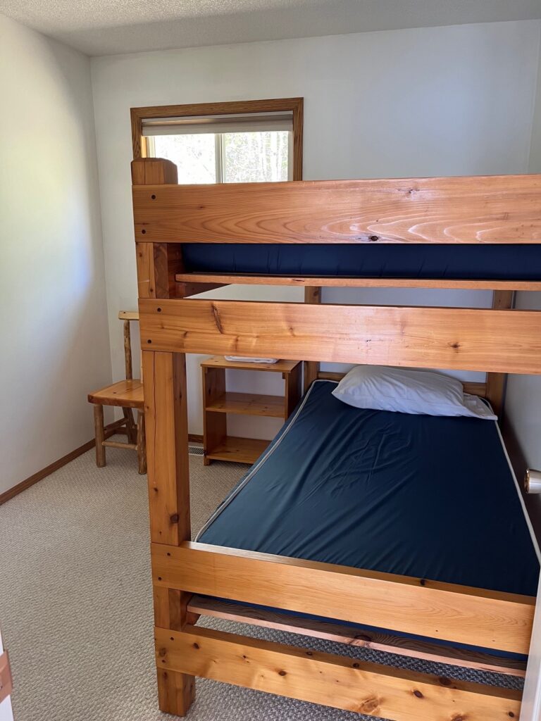 Simple room with wooden bunk bed, blue mattress, pillow, small desk, chair, and window with view of trees.