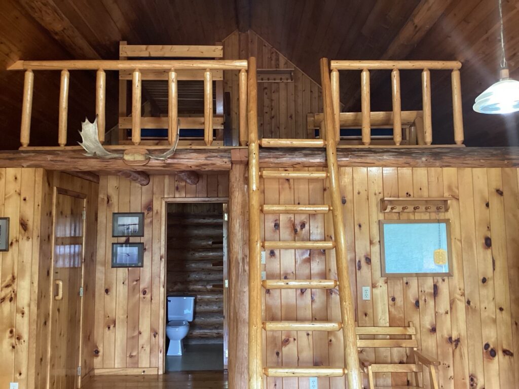 Interior of a rustic wooden cabin with a loft, ladder access, a moose antler on the railing, and visible toilet in the background.