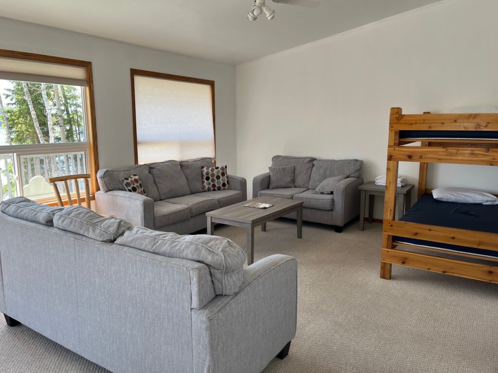 Living room with three gray sofas, a wooden bunk bed, coffee table, and large windows with a view of trees outside.