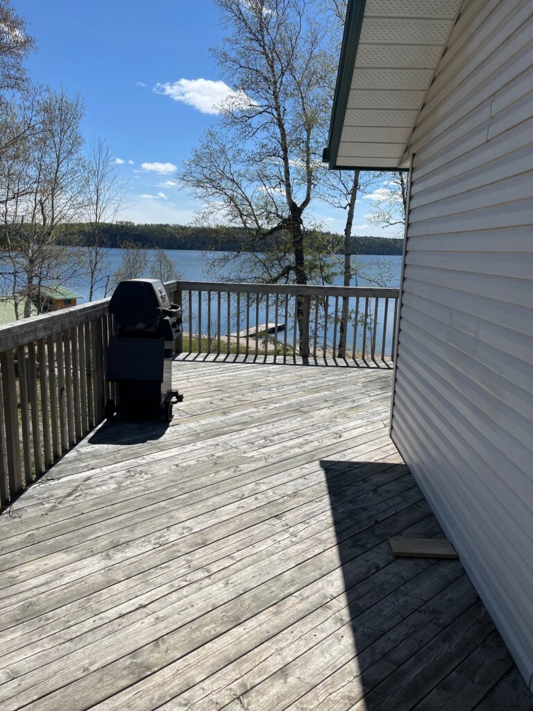 Wooden deck with a grill, railing, and view of a lake and trees under a blue sky. Side of a light-colored building visible.