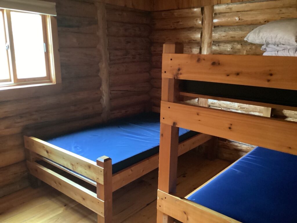 Interior of a log cabin room with wooden bunk beds and a window on the left. Blue mattresses are on the beds.