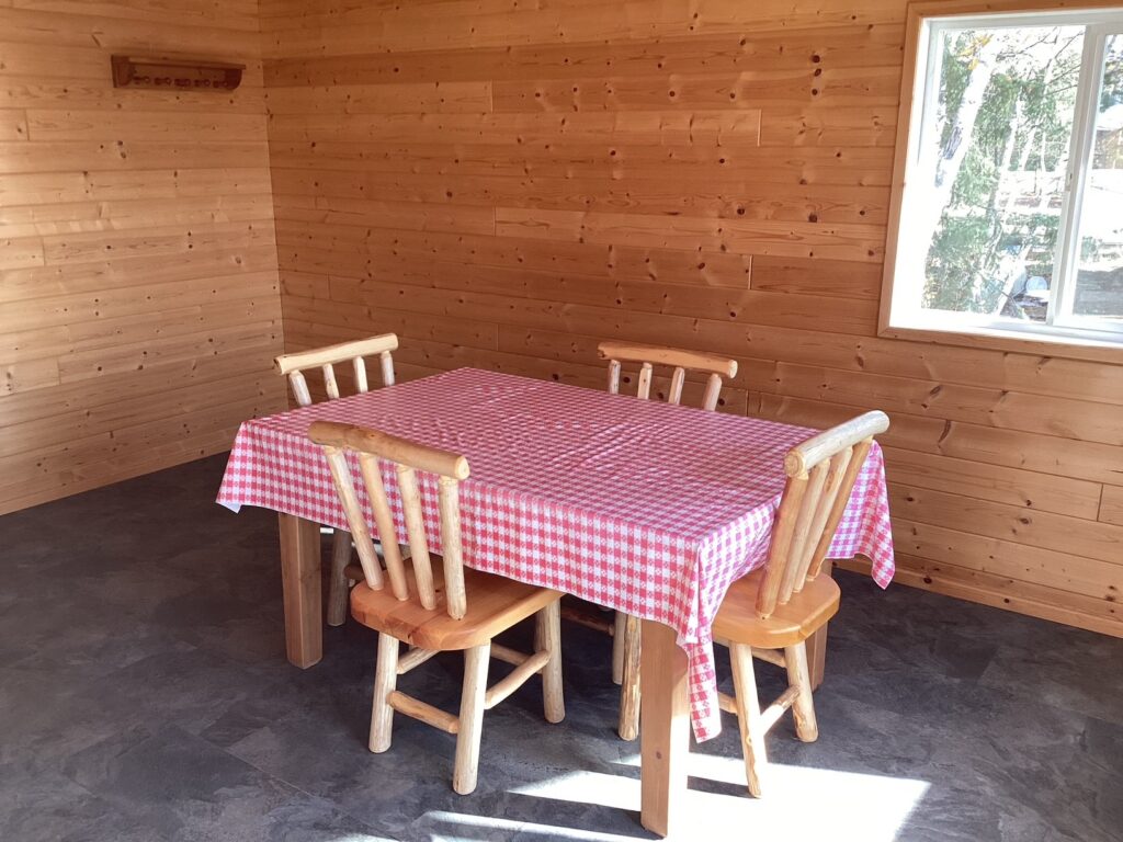 Wooden table with a red-checkered tablecloth and four matching chairs in a wooden-paneled room with a window.