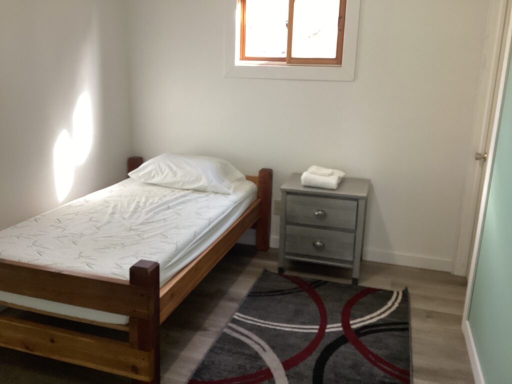 Simple bedroom with a single bed, a gray nightstand with folded towels, and a patterned rug. Small window above.