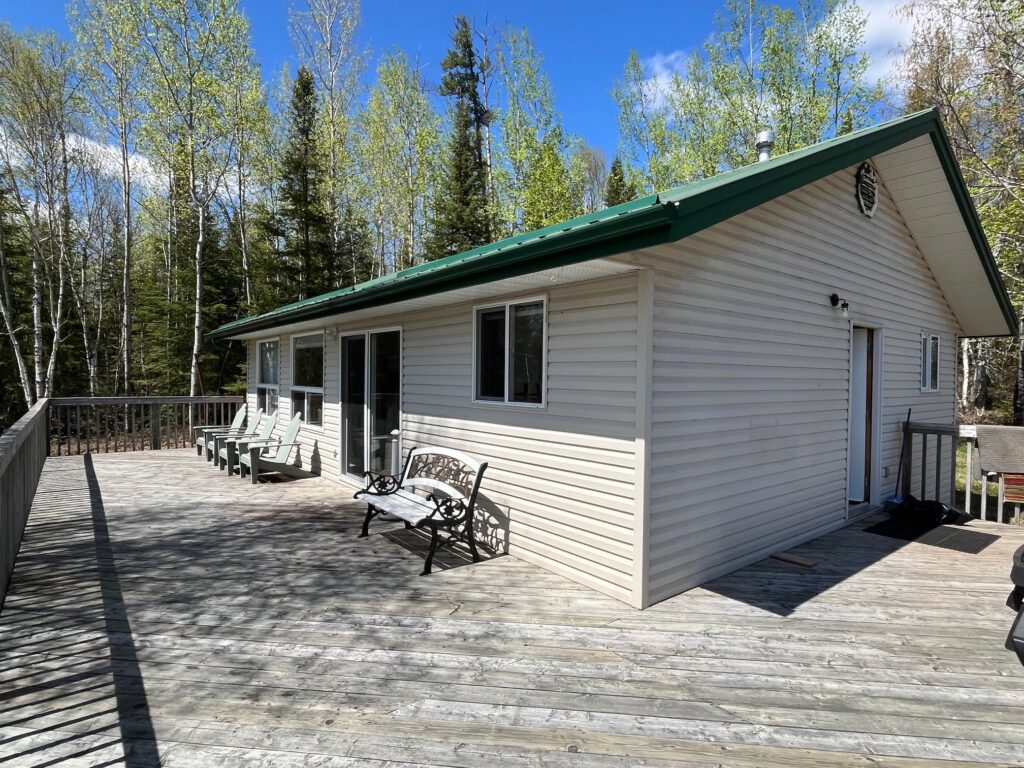 Small beige cabin with green roof on a wooden deck; surrounded by trees under a blue sky with clouds.