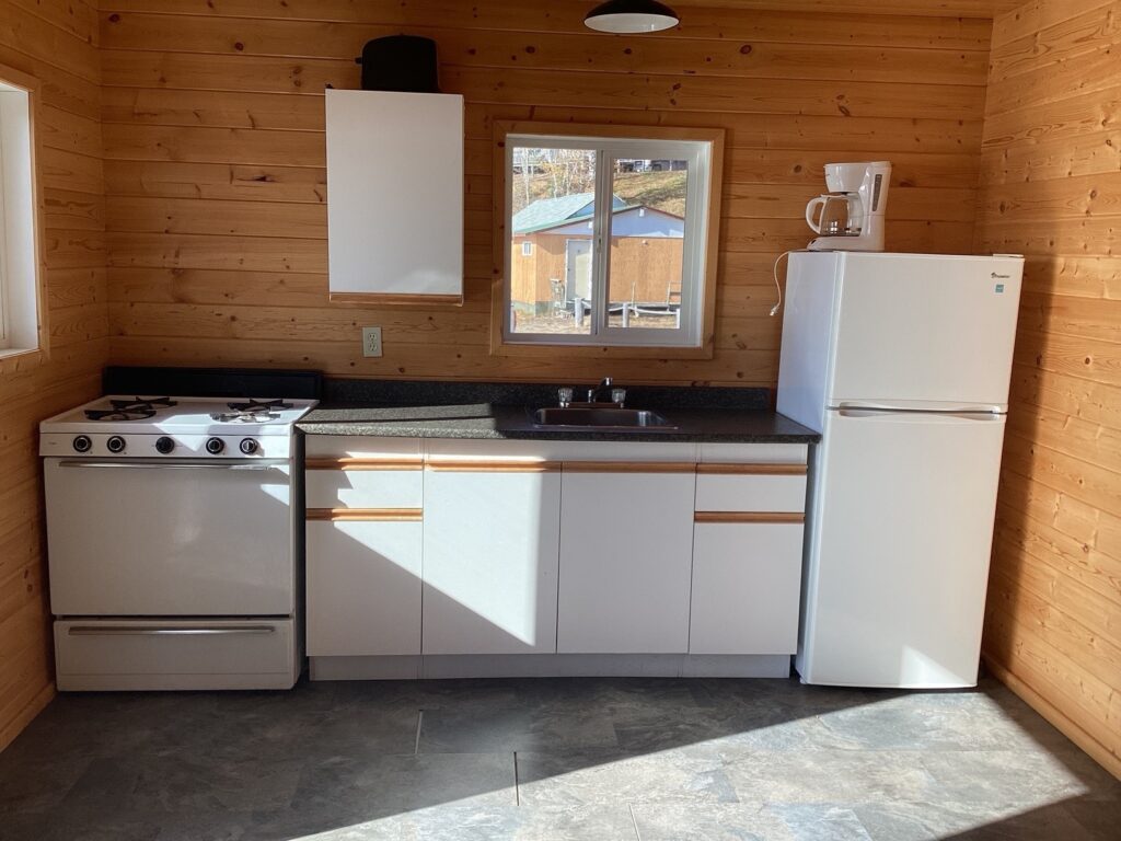 A small kitchen with wood-paneled walls features a white stove, cabinets, fridge, sink, and a coffee maker on the counter.