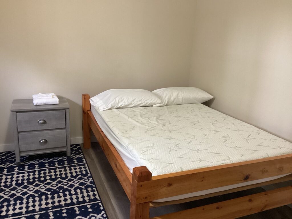 A simple bedroom with a wooden double bed, white linens, a small gray nightstand with a folded towel, and a patterned rug.