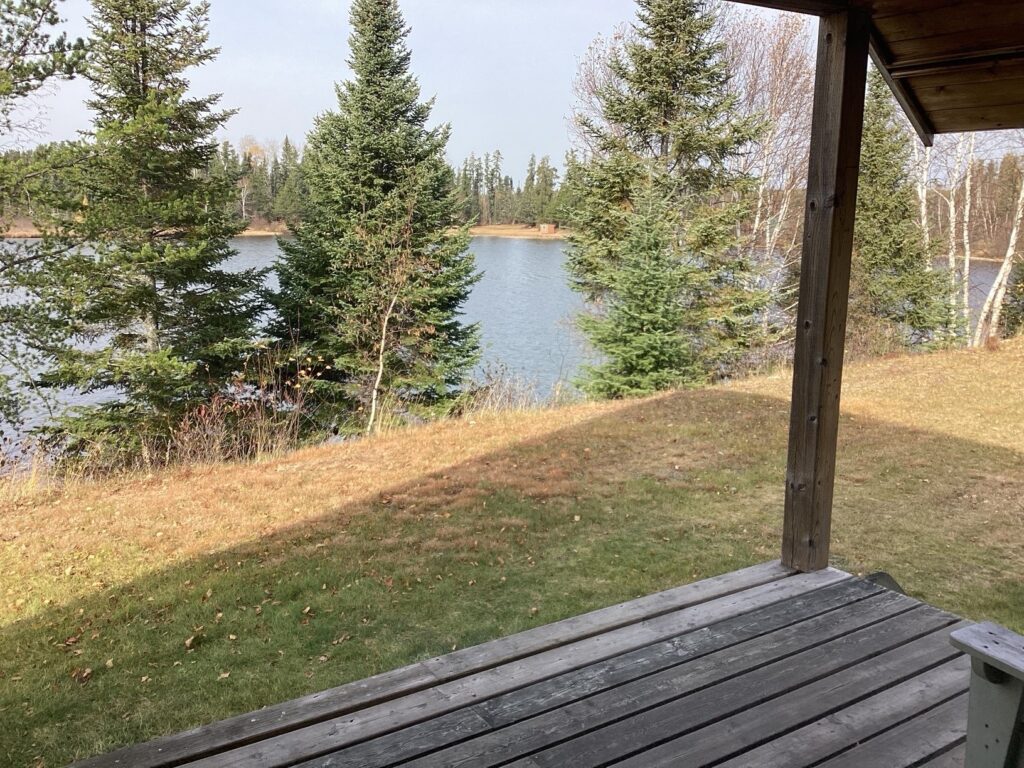 View from a wooden porch overlooking a grassy area and a serene lake, surrounded by evergreen trees and a partly cloudy sky.