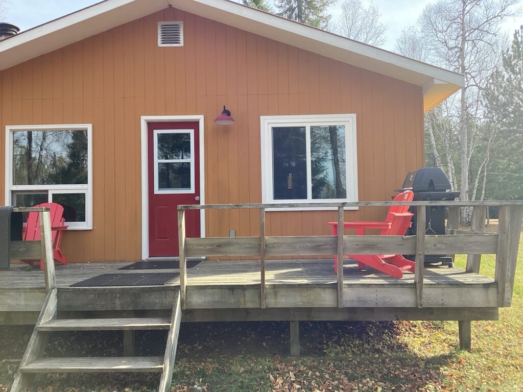 Wooden cabin with orange siding, red door, and a porch. Two red chairs and a barbecue grill are on the porch. Trees in background.