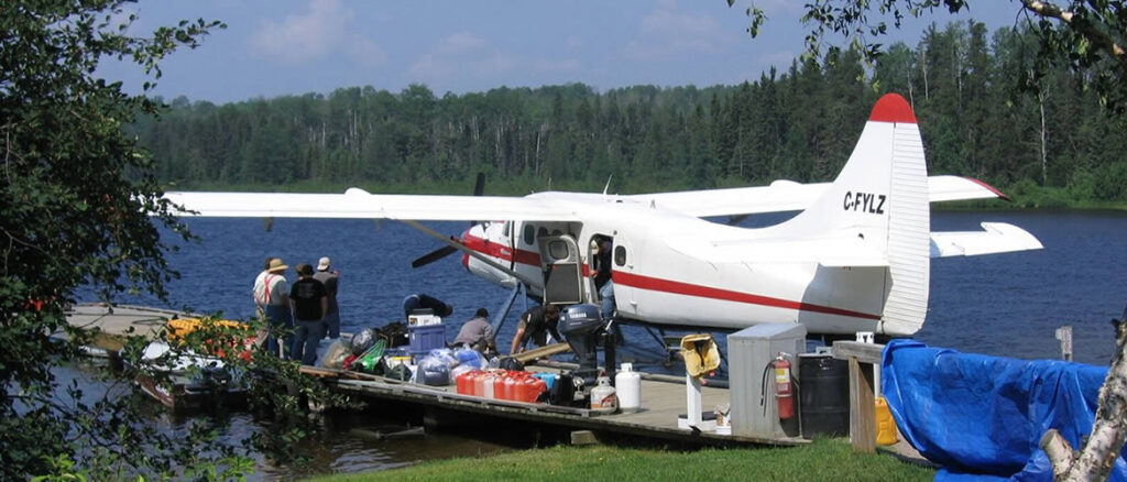 Seaplane docked by a lake with several people loading supplies onto it, surrounded by trees under a blue sky.