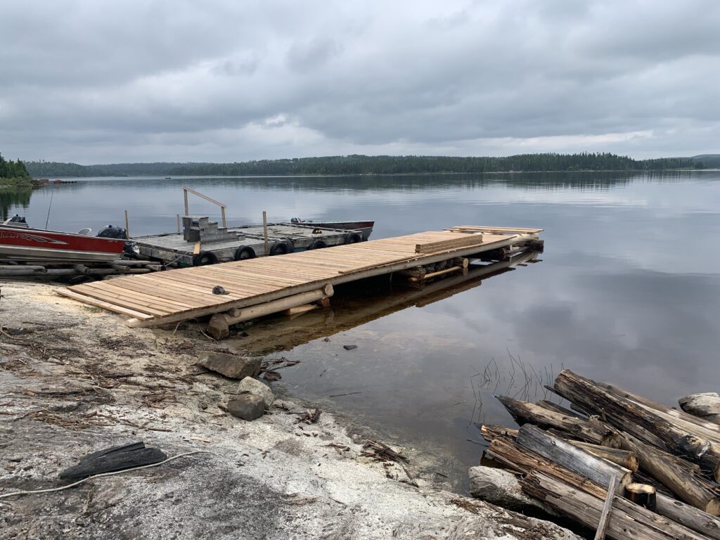 Wooden dock extending into a calm lake, with a boat tied alongside under a cloudy sky. Logs and rocks on the shore.