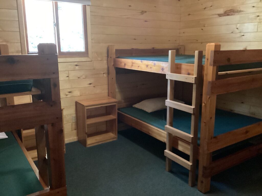 A small wooden room with two wooden bunk beds, a small window, and a bedside table. The floor is carpeted.