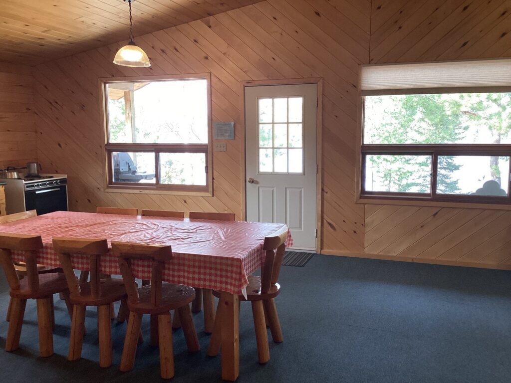 Wooden dining room with a long table covered by a red checkered cloth, six chairs, a stove, and large windows by a door.