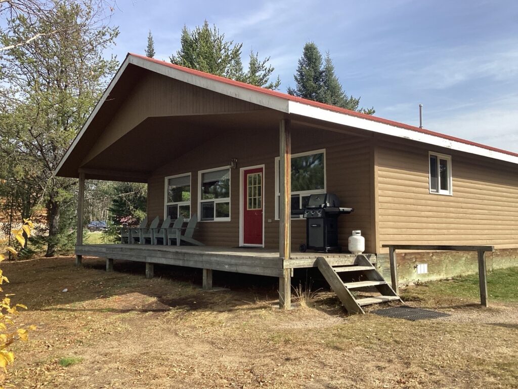 A small brown cabin with a red door, porch with chairs, and barbecue grill, surrounded by trees under a clear sky.