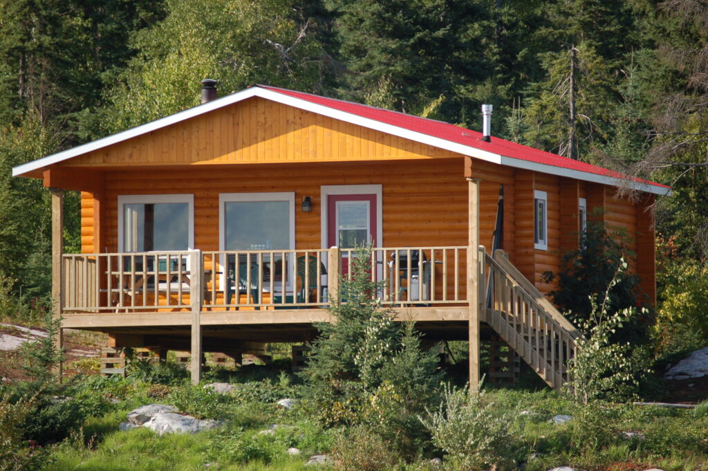 A small wooden cabin with a red roof and a porch, surrounded by trees and greenery.