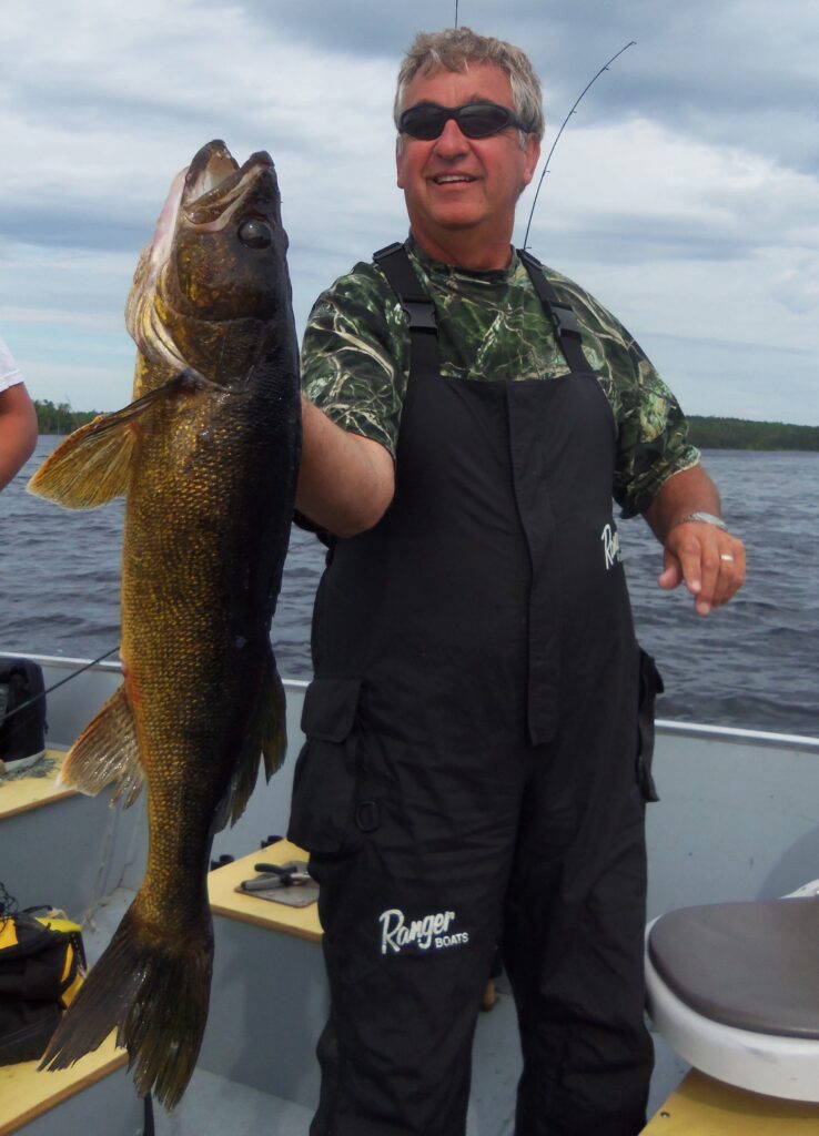 Person on a boat holding a large fish, wearing a camo shirt and black overalls, with water and an overcast sky in the background.