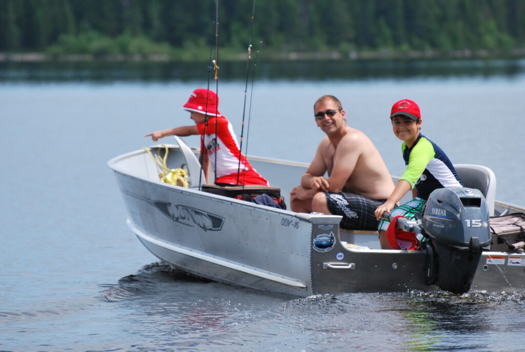 Three people on a small motorboat fishing in a lake; two children in red hats and a man shirtless. Forest visible in background.