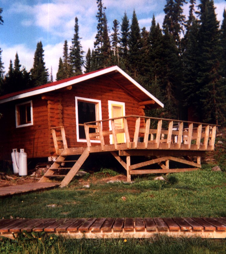 A wooden cabin with a deck is situated in a forested area. There is a path leading to the porch, surrounded by grass and trees.