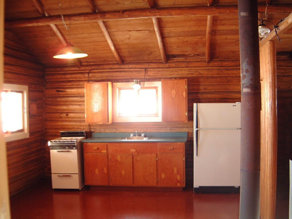 Rustic log cabin kitchen with wooden cabinets, white fridge, stove, overhead light, and exposed beam ceiling.