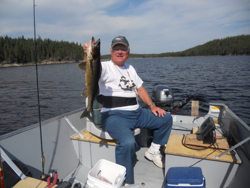 Man sitting in a boat on a lake, holding a fish he caught, with trees in the background and fishing equipment nearby.