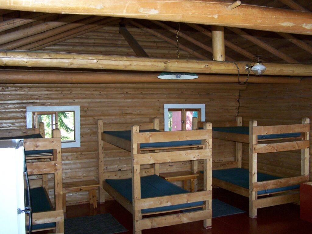 Interior of a wooden cabin with four twin bunk beds, blue mattresses, log walls, and exposed ceiling beams.