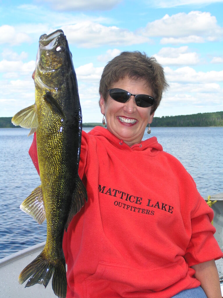 Person wearing a red hoodie and sunglasses, smiling while holding up a fish on a boat in front of a lake.