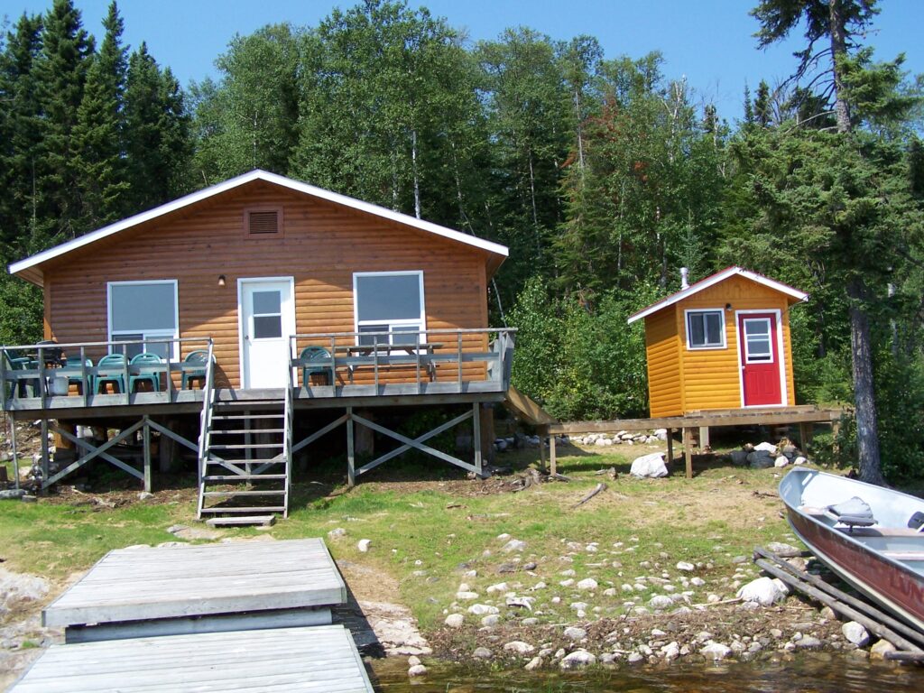 Rustic cabin with front steps and chairs on a wooden deck, next to a small shed, surrounded by trees, near a lake with a dock.