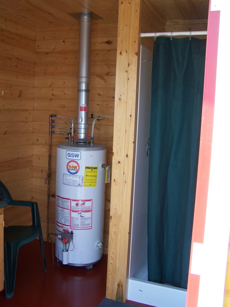 A water heater is installed in a wooden room next to a shower with a dark green curtain. A green chair is partially visible.