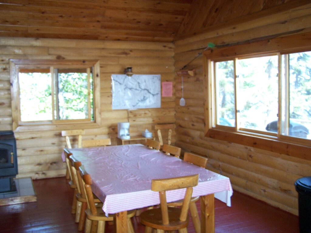 Log cabin interior with a wooden table and chairs. Red tablecloth covers the table. Windows show trees outside. Wood stove in corner.