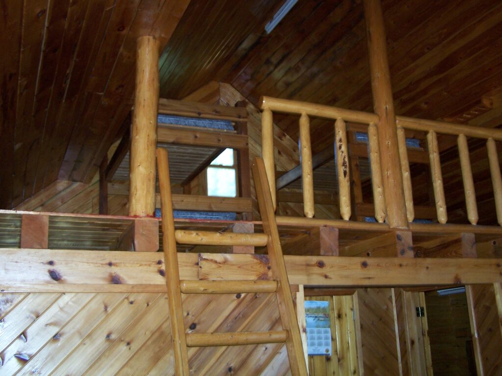 Wooden loft interior with a ladder leading to the upper level and railings; walls and ceiling made of smooth, polished wood.