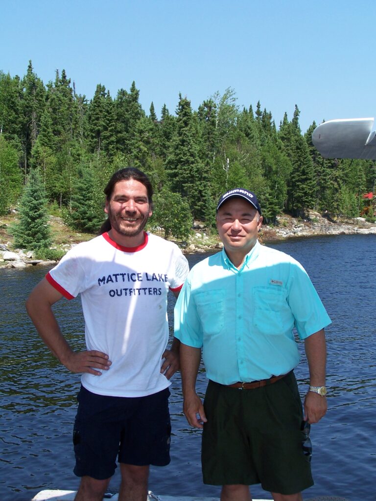 Two men stand by a lake, one in a white shirt and shorts, the other in a blue shirt and cap, with trees in the background.