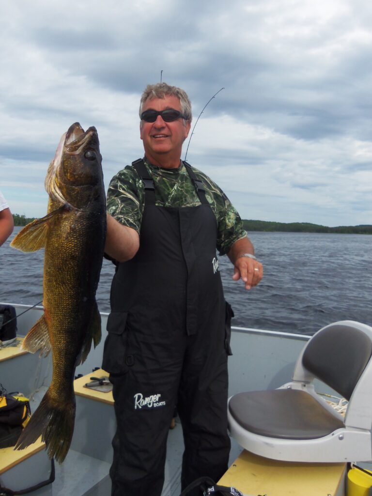 Person holding a large fish on a boat, smiling. Wearing camo shirt and black overalls under a cloudy sky.