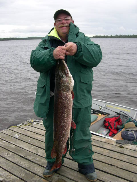 Person in green gear holding a large fish on a dock beside a boat, with a lake and cloudy sky in the background.