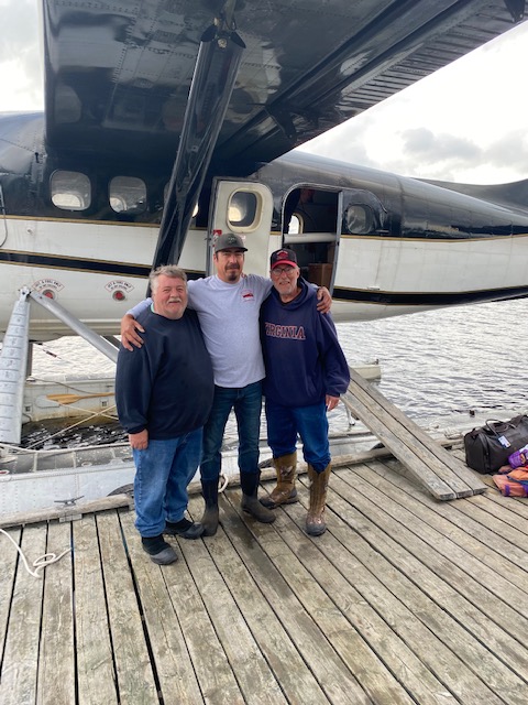 Three men standing on a wooden dock in front of a seaplane, with water and cloudy sky in the background.