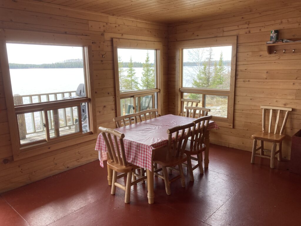 A cozy wooden dining area with a table and six chairs, surrounded by windows showing a lakeside view and trees outside.