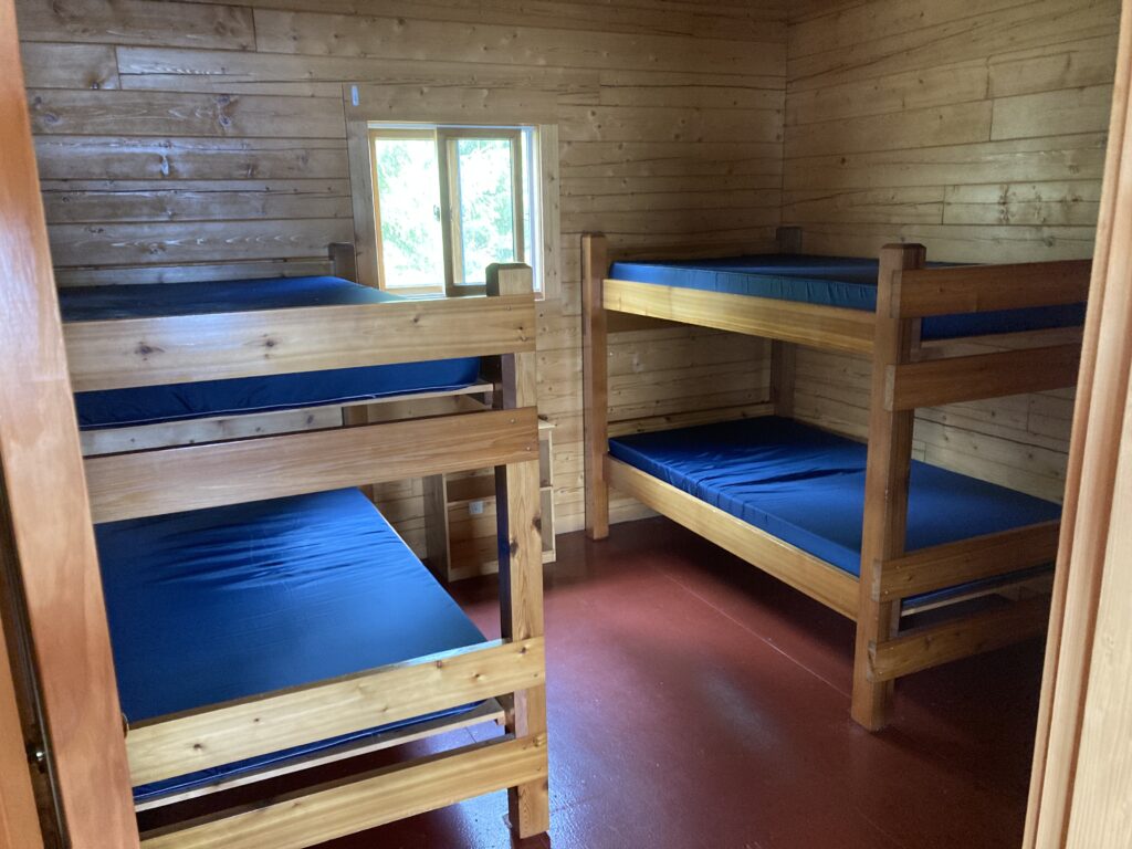 Small wooden room with two sets of bunk beds, blue mattresses, wooden walls, a window, and red-brown flooring.