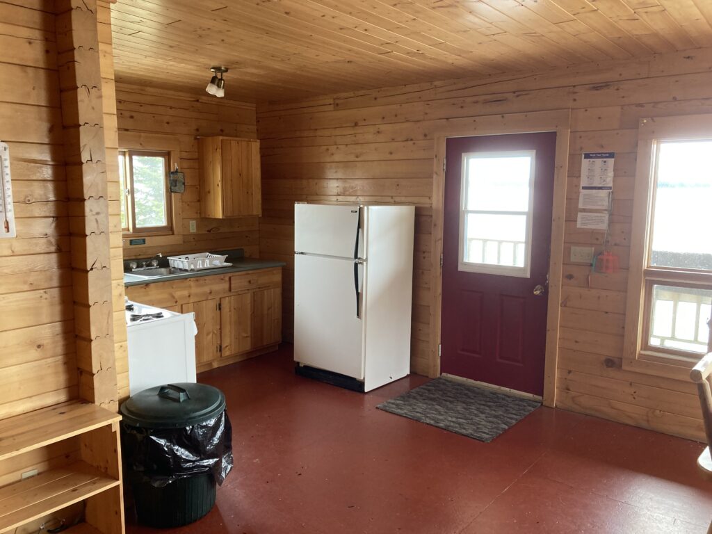 Wood-paneled kitchen with white appliances, red door, window with lake view, brown floor, and a trash bin with a black liner.