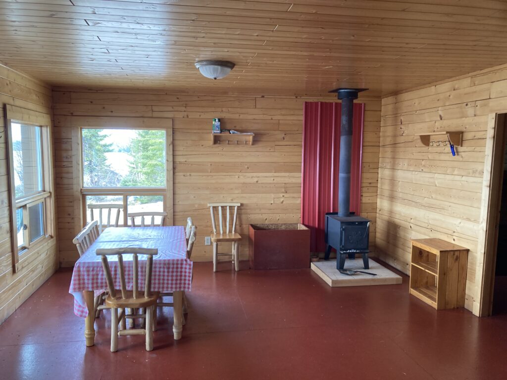 Wooden cabin interior with dining table, stove, shelves, and wide windows overlooking trees. Red curtain by the stove.