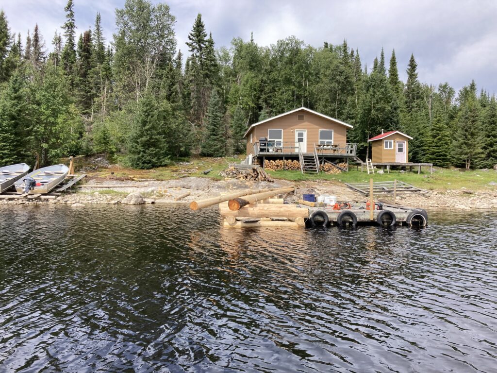 Log cabin with dock on a forested lakeshore, featuring boats on land and a small floating platform in the water.