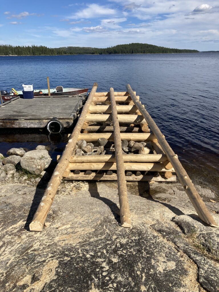 Wooden ramp and scattered rocks leading to a small dock on a lake with forested shoreline in the background.